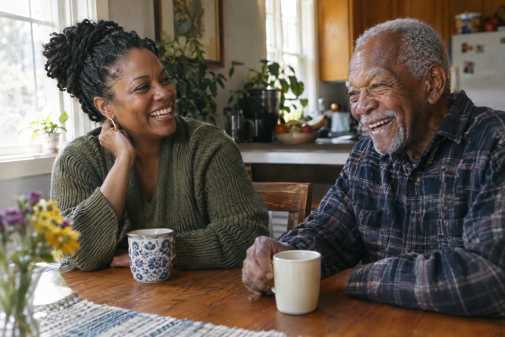A woman and an older man laughing together over coffee at a sunlit kitchen table.