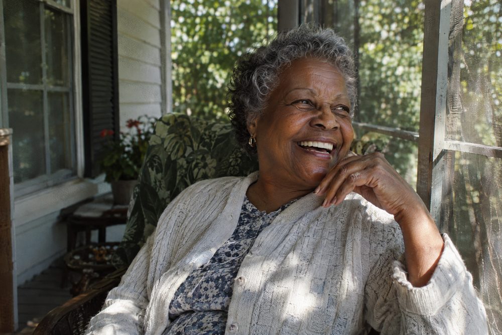 An older woman laughing on a covered front porch in summer sunlight.