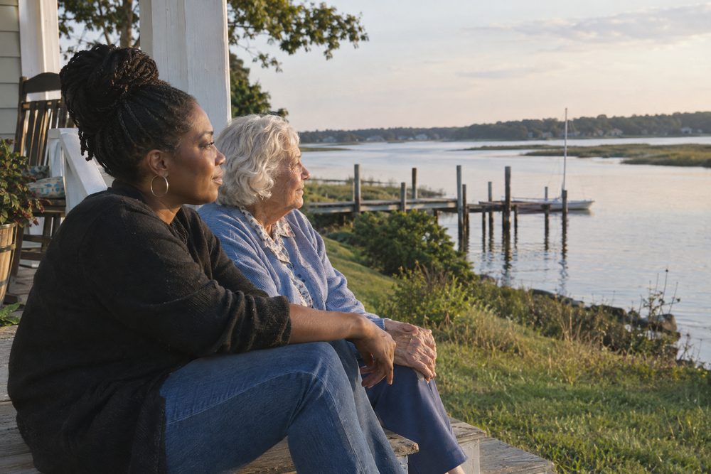 Two women sitting together on a porch step looking out at a tidal river and a dock at dusk.